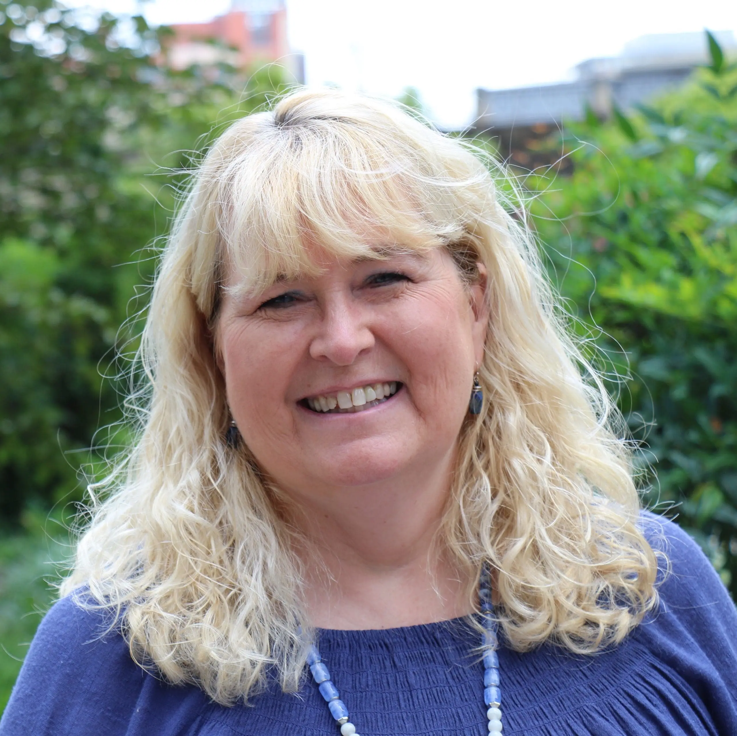 Marcia Baldwin is a white woman with light blonde hair. She is smiling for the photo, taken outdoors with greenery in the background.