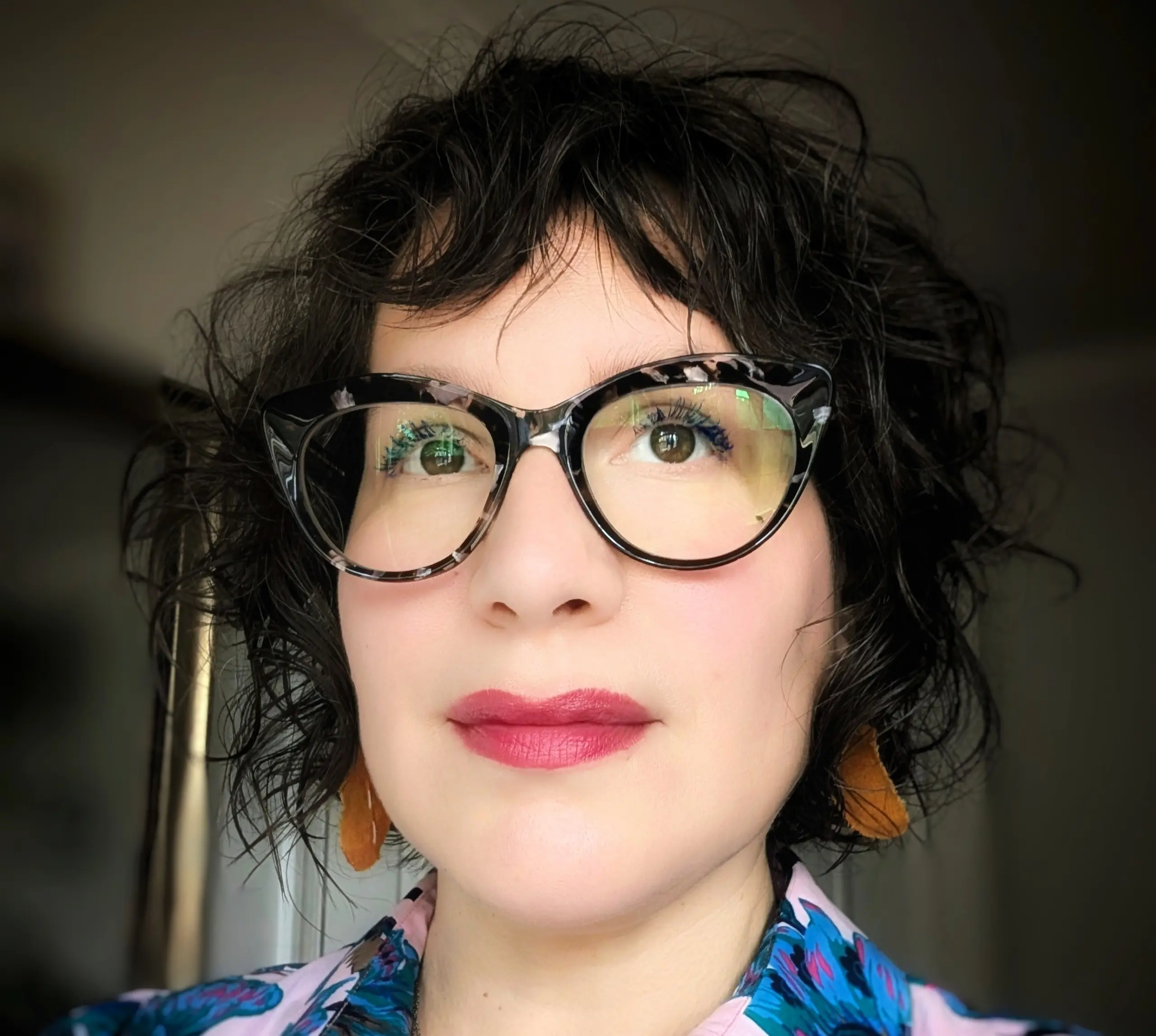 A Latina with short brown hair, wearing tortoiseshell cat-eyeglasses, earrings, & a blue & pink dress