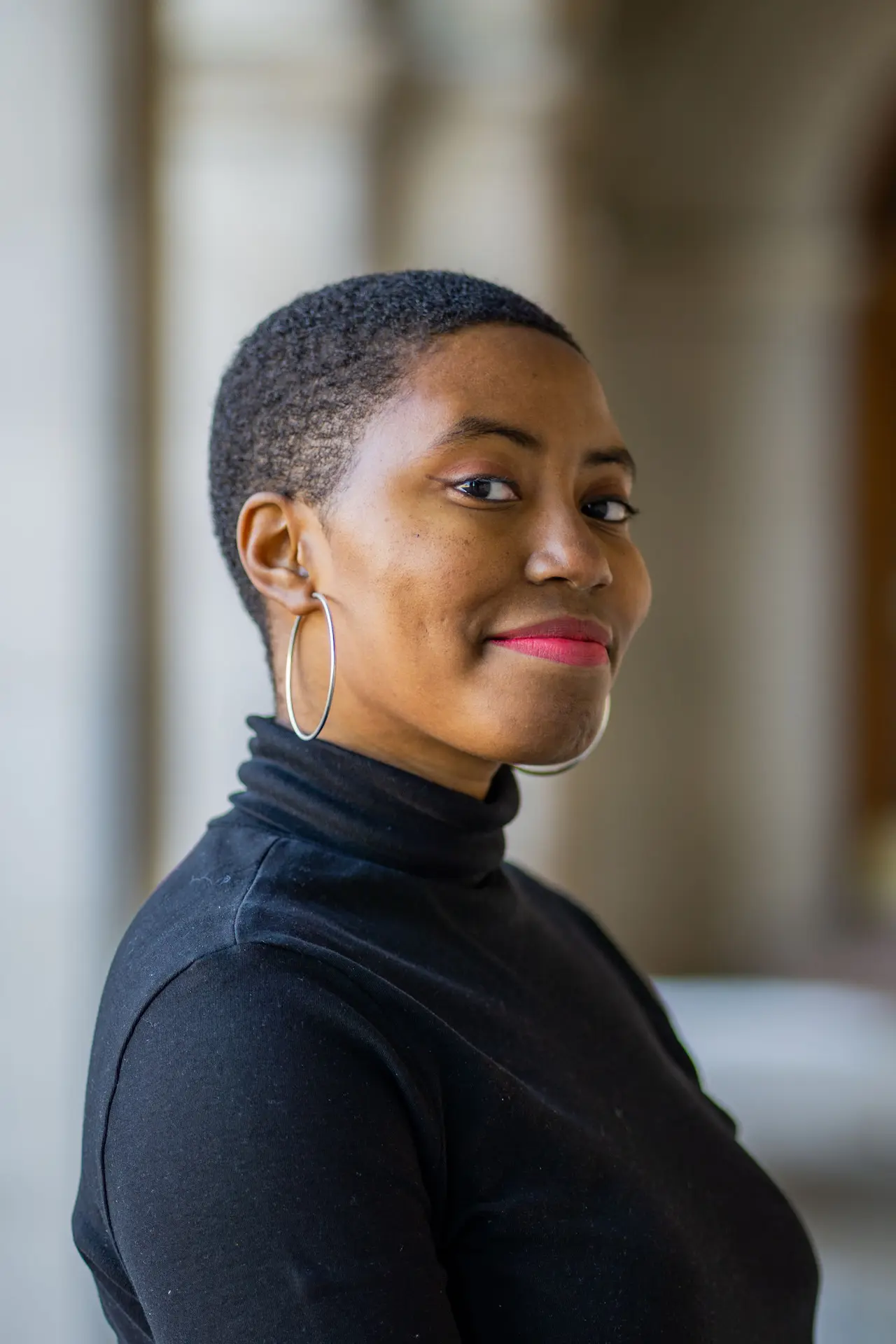 Olivia Williams, a young Black woman with low cut coily black hair wearing a black turtleneck, silver hoop earrings, and red lipstick. She is smiling softly at the camera.