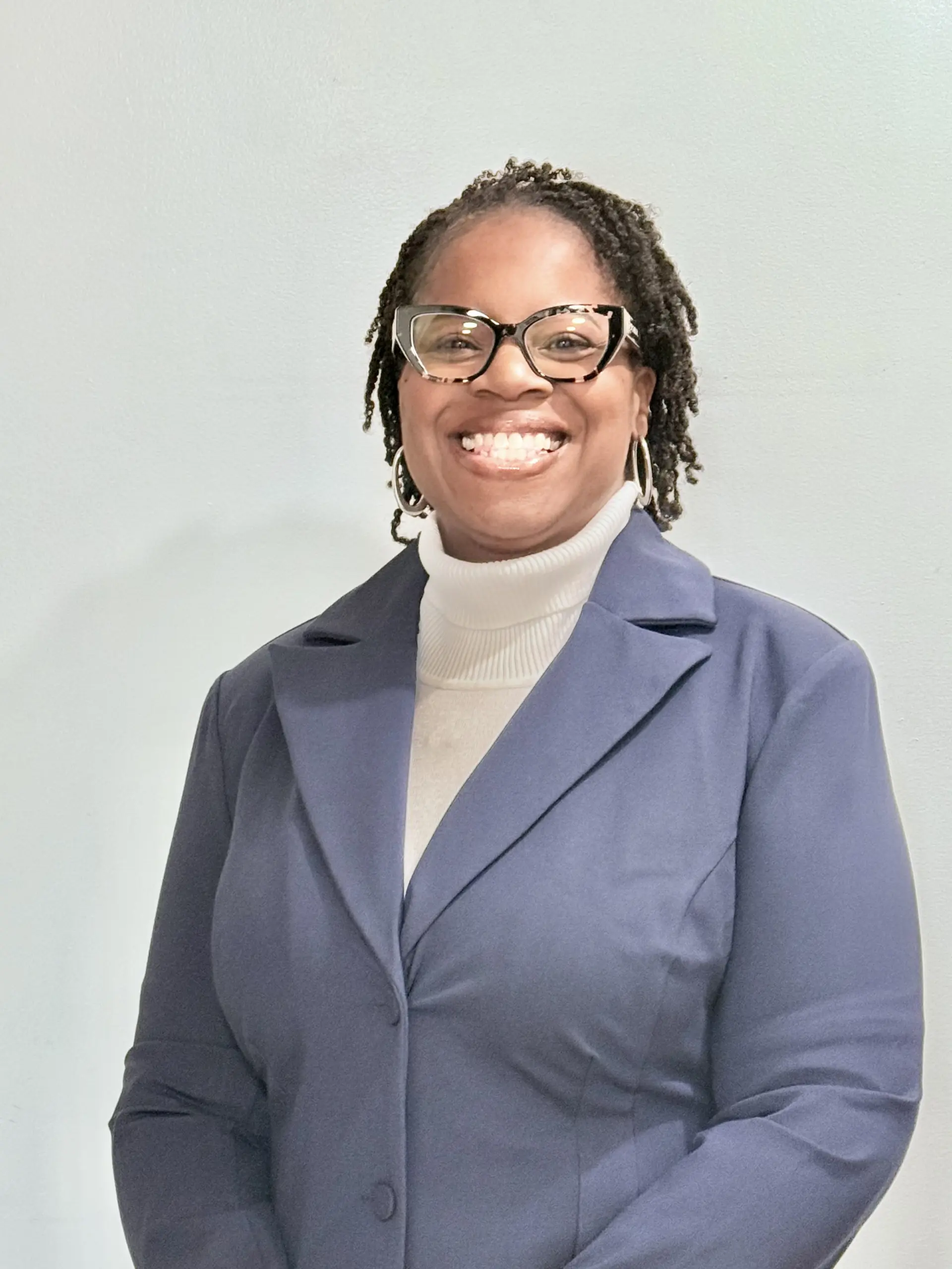 Crystal Smith-Hunt, a black woman with short twists in her hair and wearing glasses. This is a headshot, and she is wearing a white sweater and a navy-blue jacket.