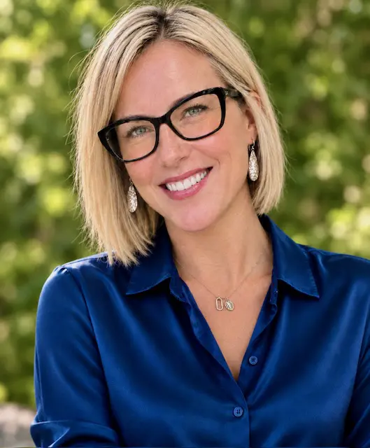 Kristin Priddy, a white woman with shoulder length blonde hair and black glasses smiling at the camera. She is wearing a blue blouse, silver earnings, and necklace.