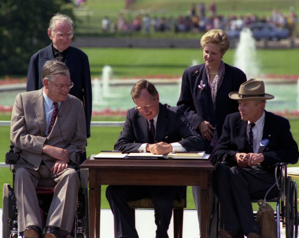 President Bush signs the Americans with Disabilities Act on the South Lawn of the White House. Sharing the dais with the President and he signs the Act are (standing left to right): Rev. Harold Wilkie of Clairmont, California; Sandra Parrino, National Council on Disability; (seated left to right): Evan Kemp, Chairman, Equal Opportunity Commission; and Justin Dart, Presidential Commission on Employment of People with Disabilities. Mrs. Bush and Vice President Quayle participate in the Ceremony. 26 July 1990 Photo credit: George Bush Presidential Library and Museum