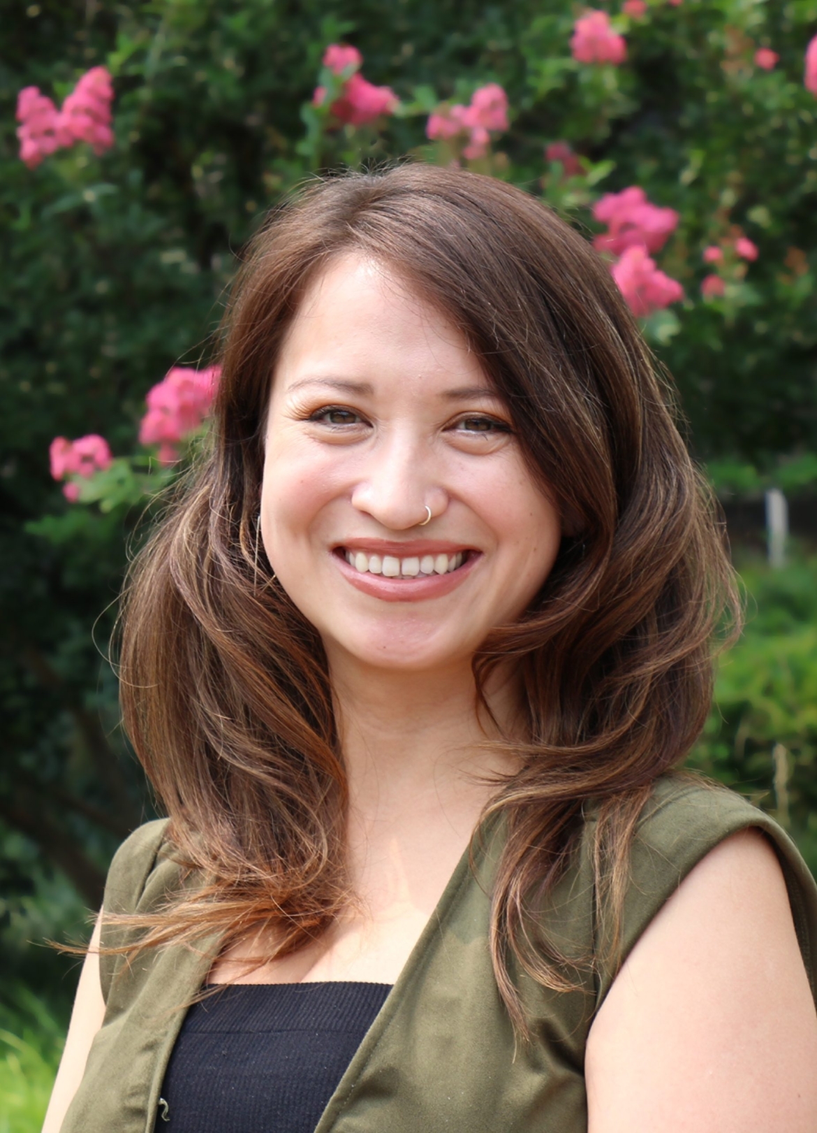 Danielle, a mixed Asian woman with long brown hair, wearing a green a black dress in front of green bushes with pink flowers