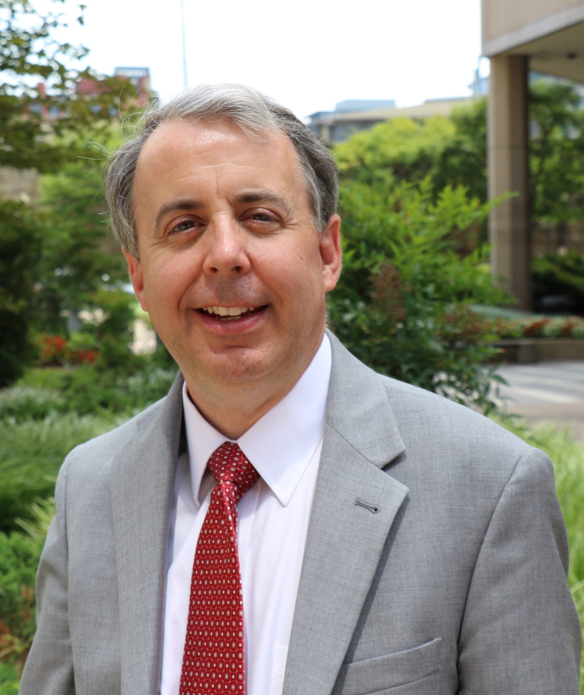 Portrait of David Hutt. He is a white man with silver and grey hair wearing a light grey jacket, white collared shirt and red tie. David is smiling at the camera, in front of a background of greenery.