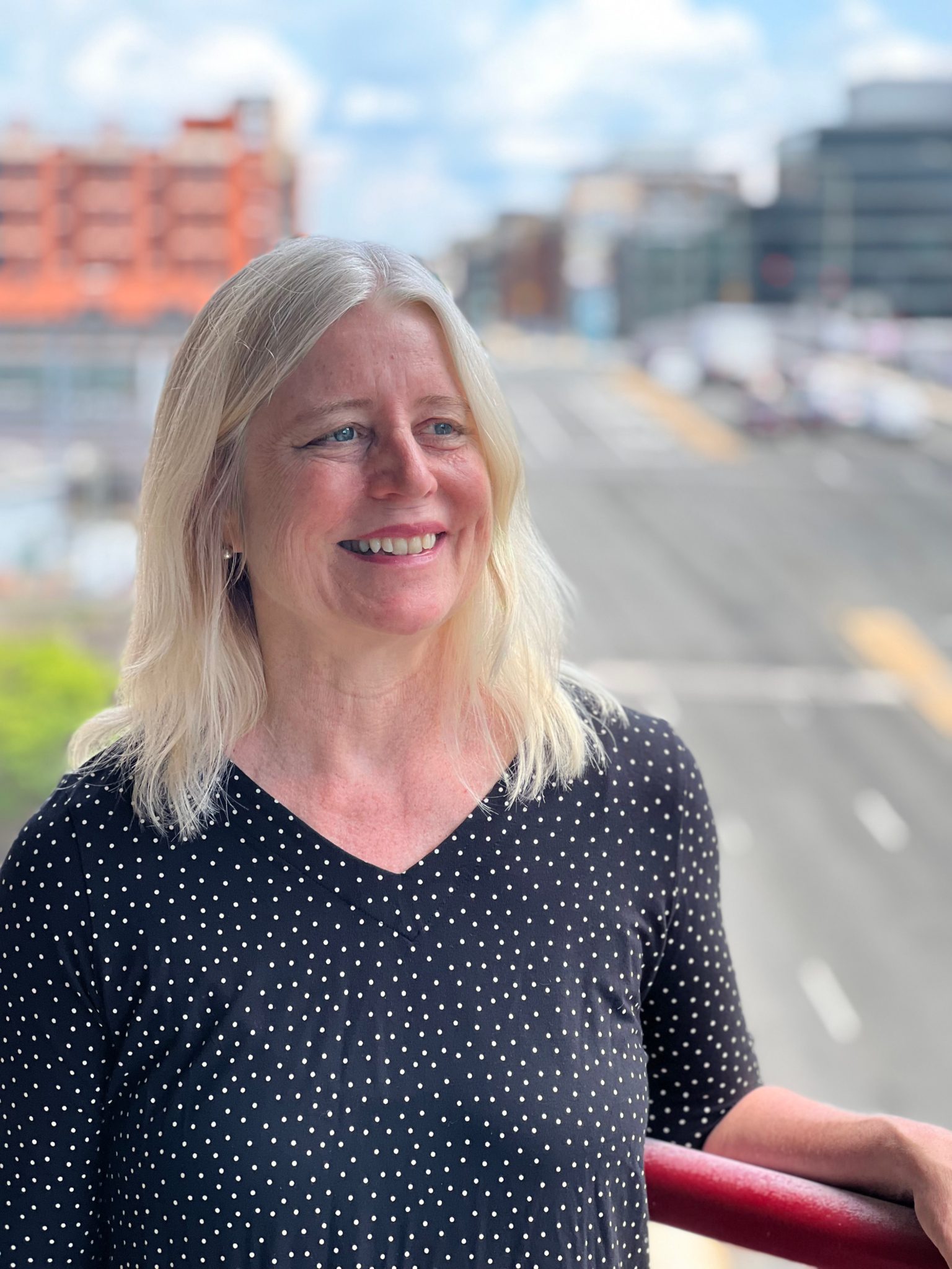 Portrait of Diane Smith Howard. Diane is a white woman with light blonde hair and blue eyes. She is black shirt with white dots on it. She's standing on a balcony, with a DC street in the background and high rise buildings. The background is blurred.