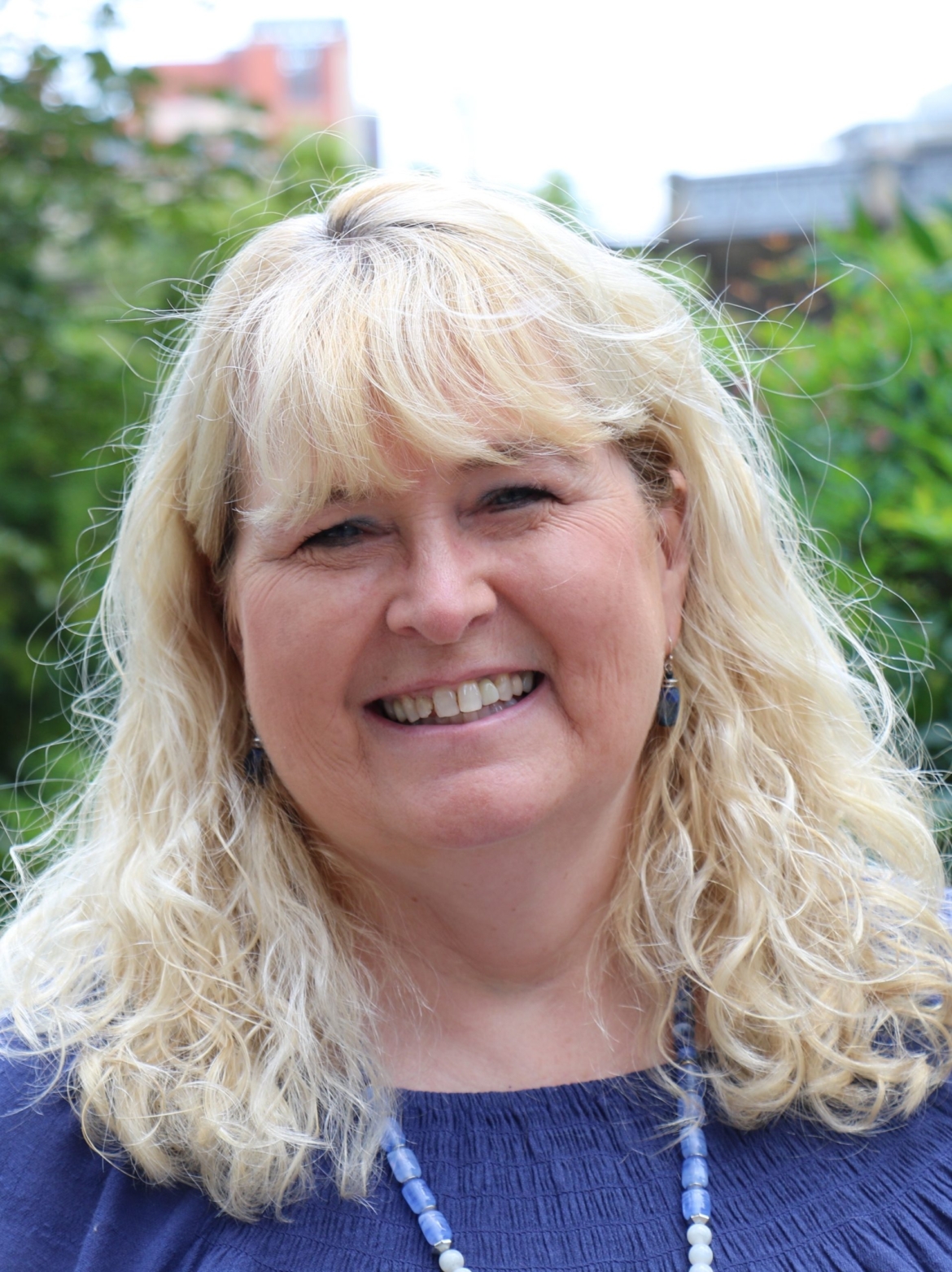 Marcia Baldwin is a white woman with light blonde hair. She is smiling for the photo, taken outdoors with greenery in the background.