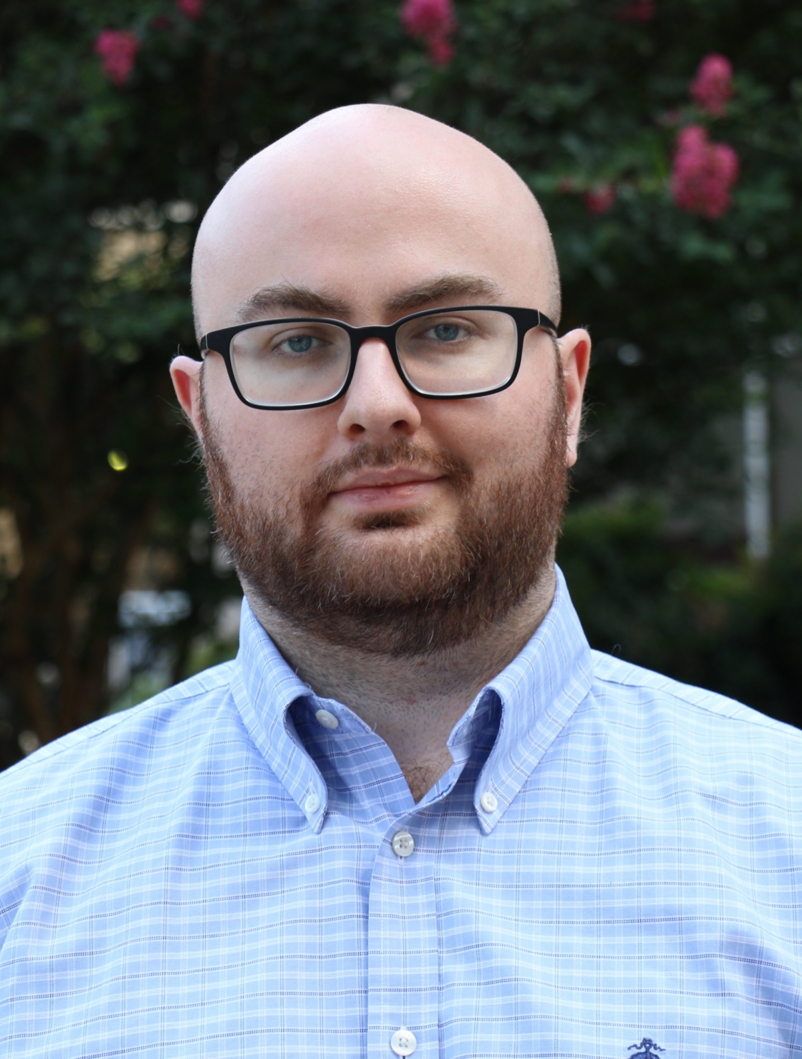 Milo is a white man with a shaved head and brown beard. He's wearing glasses and a blue button down shirt