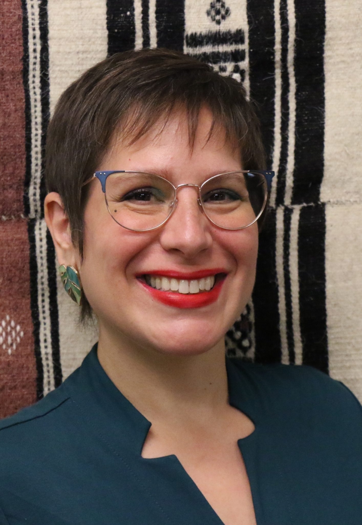 Raquel is smiling towards the camera. She is Latina with short brown hair, glasses, & a dark green dress. She is standing in front of a handwoven tapestry with geometric designs