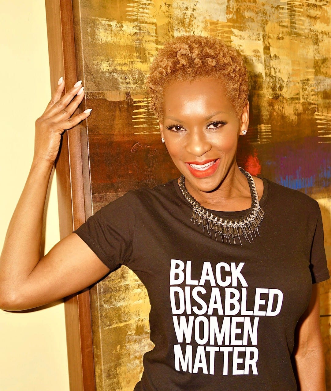 Claudia smiling looking into the camera wearing a black t-shirt with white letters that reads “Black Disabled Women Matter”