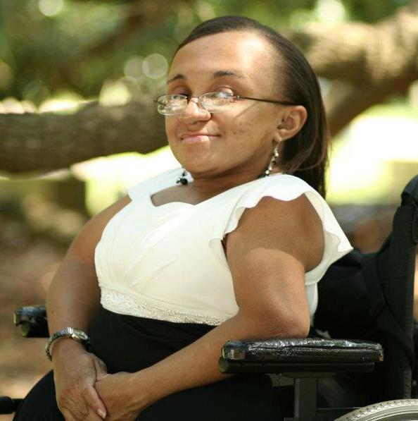 Vilissa looks to the camera over her shoulder. She wears a white top and sits in a black wheelchair. 