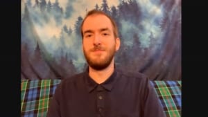 Christopher Hayes, a white male in his 20s with light brown heair and a beard, smiles directly at the camera. He is sitting infront of a tapestry hanging with forrest trees.