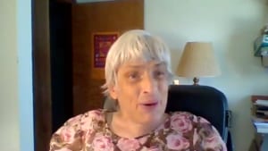 Mary-Ann, an older woman with white hair, wears a pink top and looks to the camera