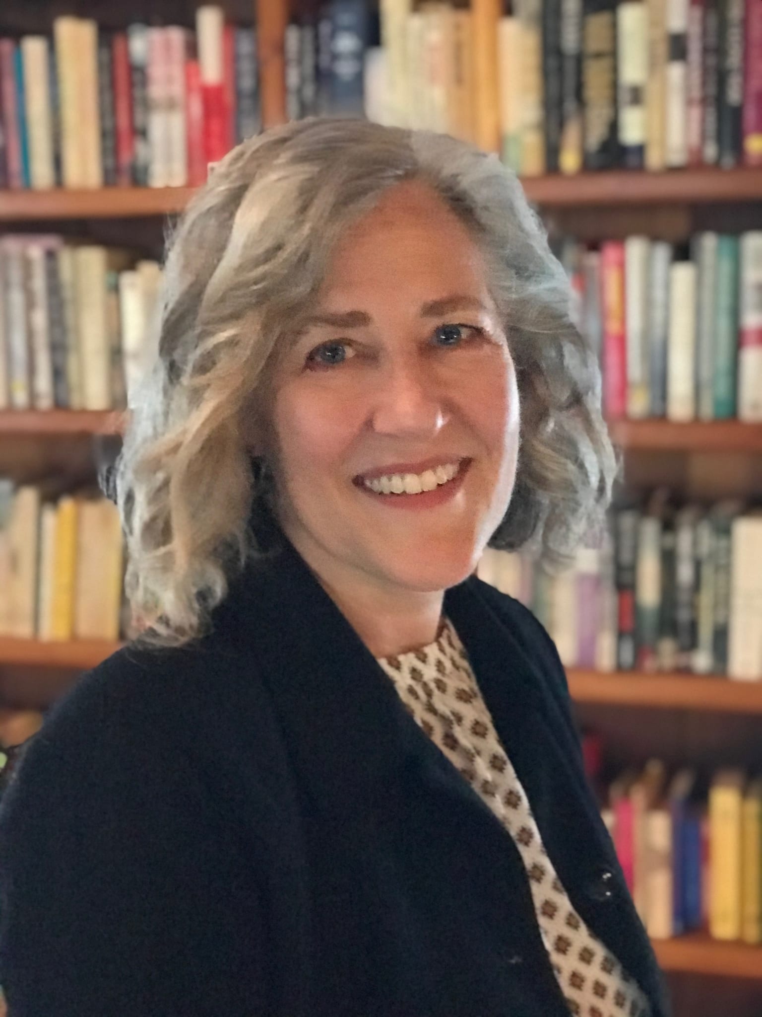 Portrait of Elissa, a middle-aged whit woman with shoulder-length, wavy, gray hair wearing a black jacket over a white print shirt, in front of blurred bookcase.