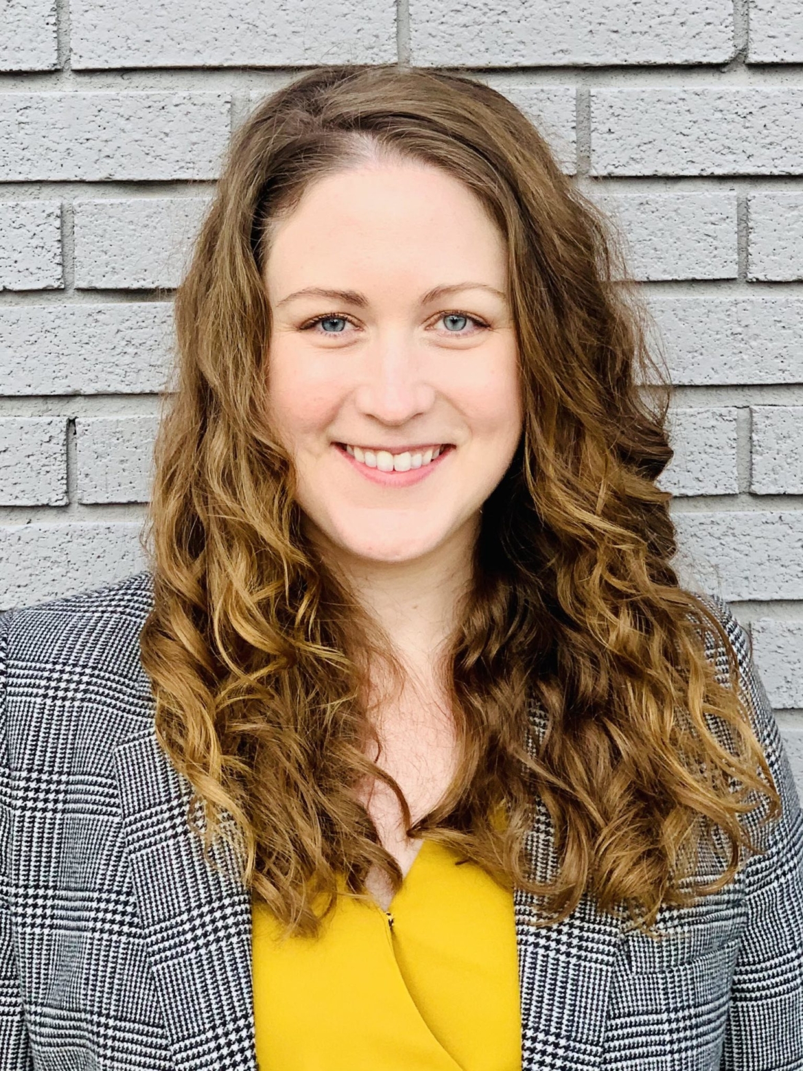 Devon is a white woman in her thirties with long, light brown curly hair. She is wearing a yellow top with a gray plaid blazer and is standing in front of a light gray brick wall.