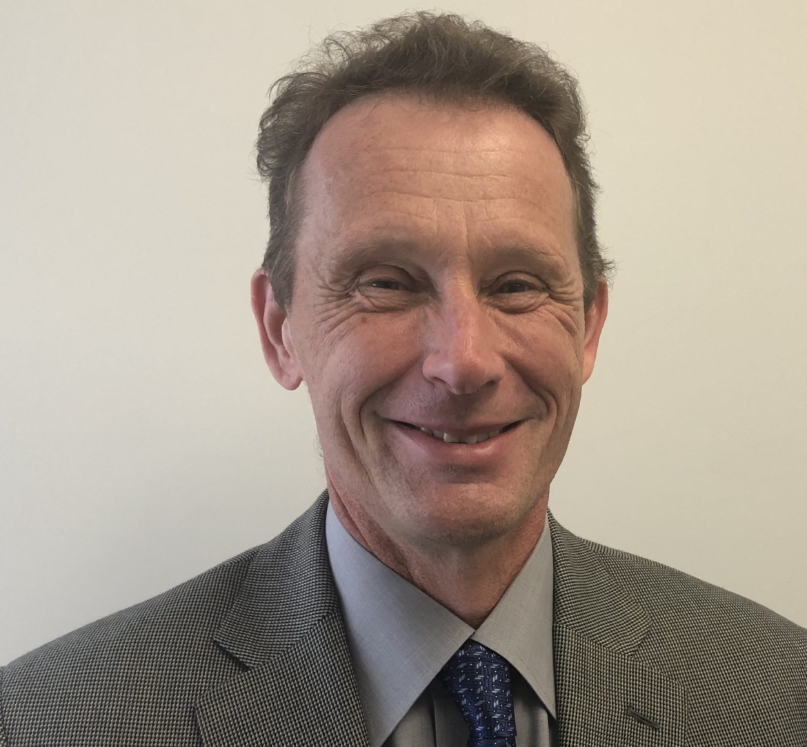 Photo of dan. He is a white man with short brown hair. He's smiling for the camera. He's wearing a houndstooth grey and white suit jacket, a light grey collared shirt, and tie with a dark and light blue pattern. Background is a nondescript off-white wall.