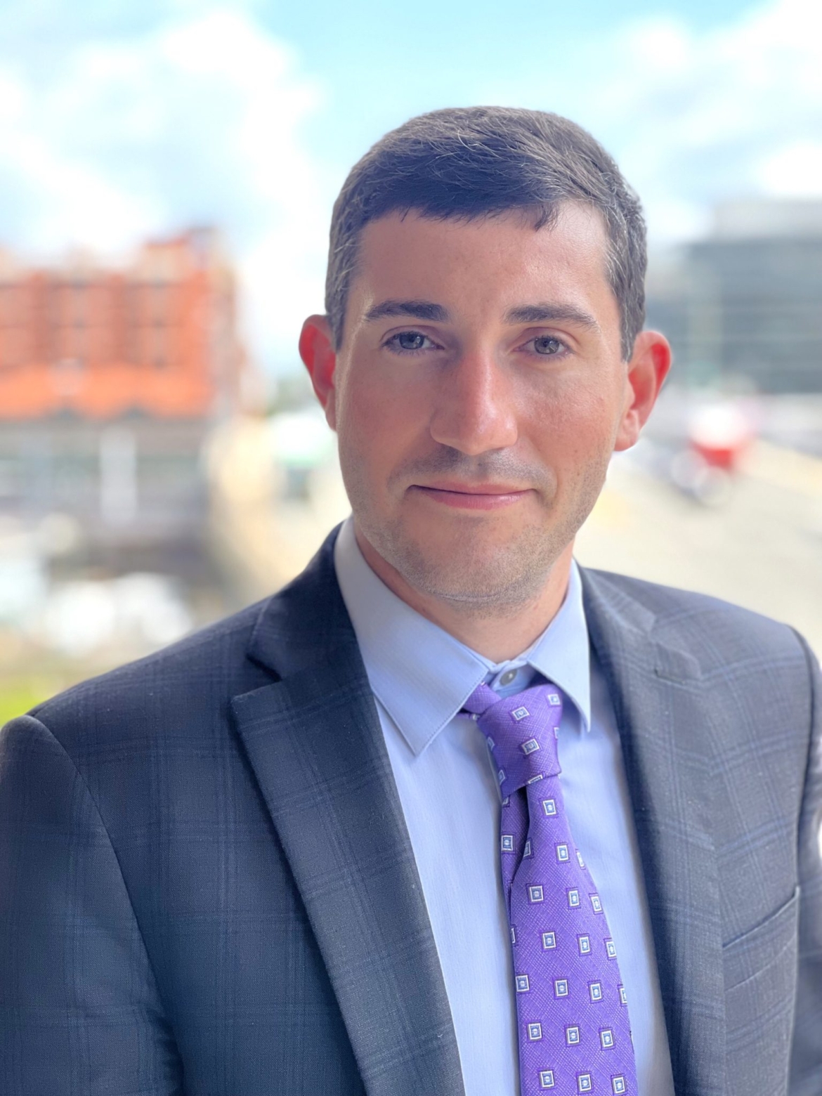Portrait of Cory Bernstein. Cory is a white man with dark hair and brown eyes wearing a dark gray suit with a faint purple pattern, a blue dress shirt, and a purple tie patterned with silver and blue squares. He is standing outside behind a blurred background on a sunny, but slightly cloudy, day.