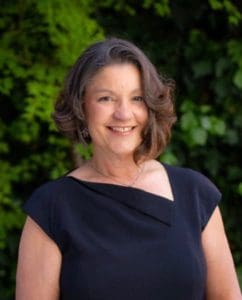 Portrait of Marlene Sallo, a Latina with brown hair, wearing a black dress. She is standing outside with greenery in the background.