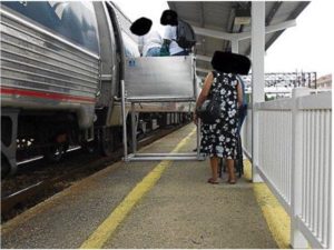 People using a wheelchair lift to board an Amtrak train. There faces are covered to protect privacy. 