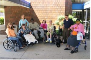 A diverse group of rail passengers with disabilities, seated outside a train station on a sunny day.