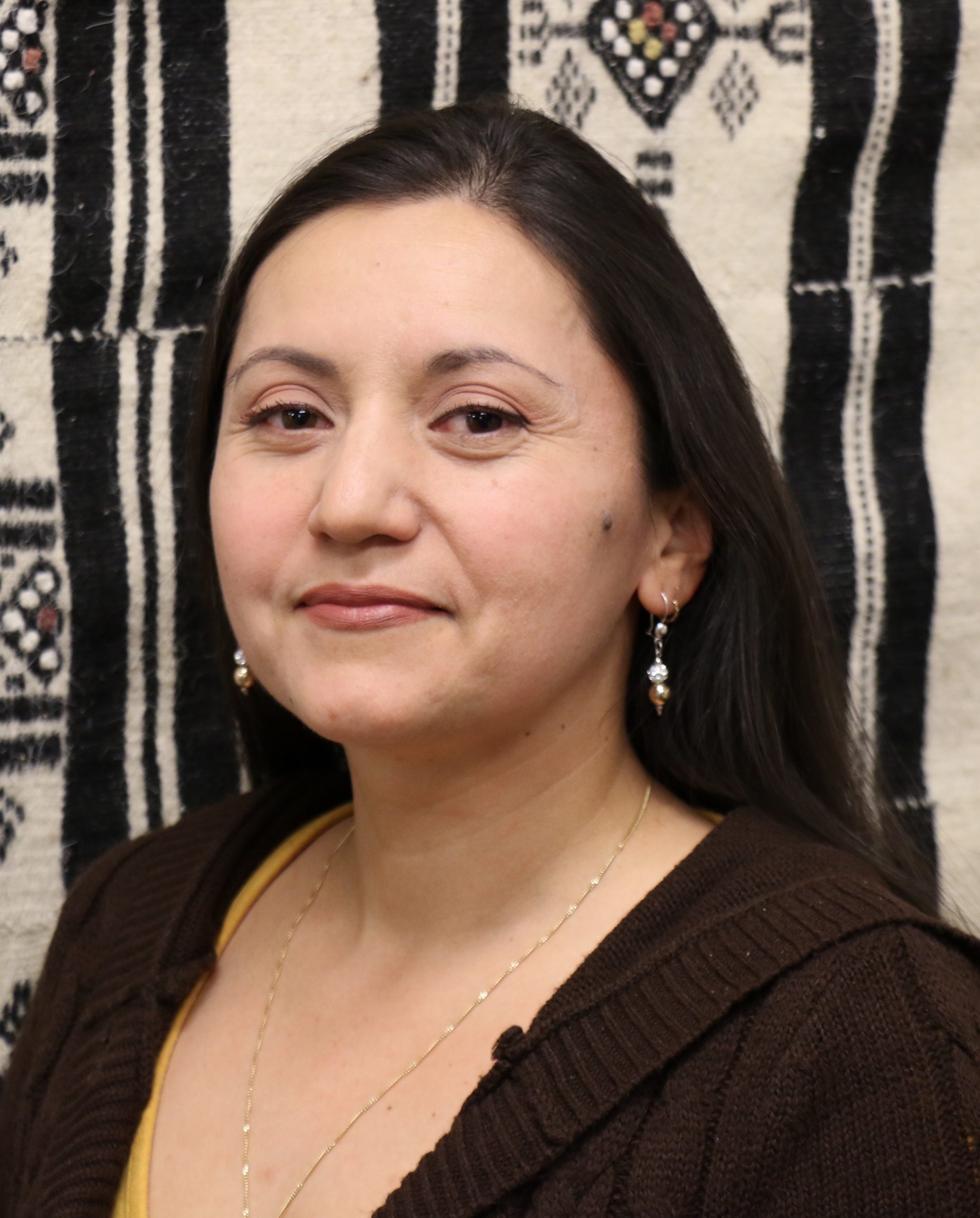 Hilda is a Colombian woman with long dark hair and brown eyes. She is wearing a brown sweater and earrings. She is standing in front of a black and white tapestry.