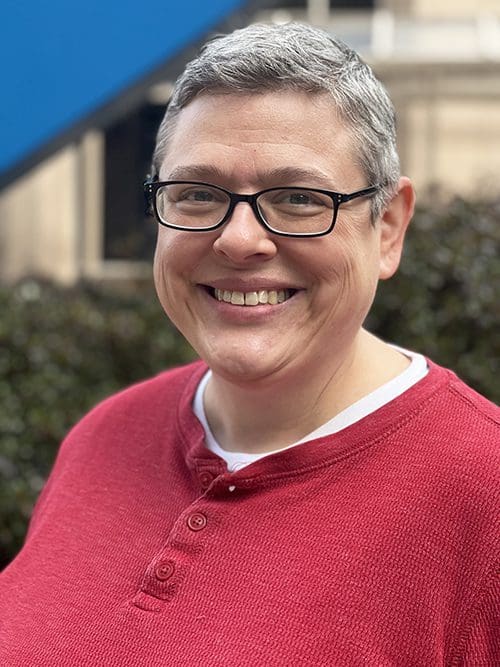 Portrait of Elise, a smiling woman with very short gray hair, wearing glasses and a red shirt.