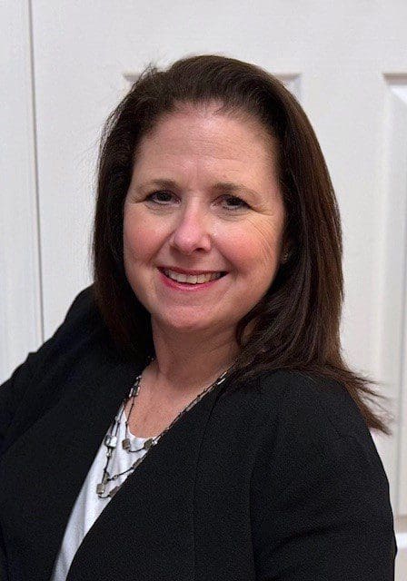 White female with brown shoulder length hair wearing a white blouse and black jacket.