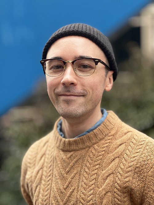 A photo of a white male wearing a brown sweater over a blue button up with glasses and a black beanie.