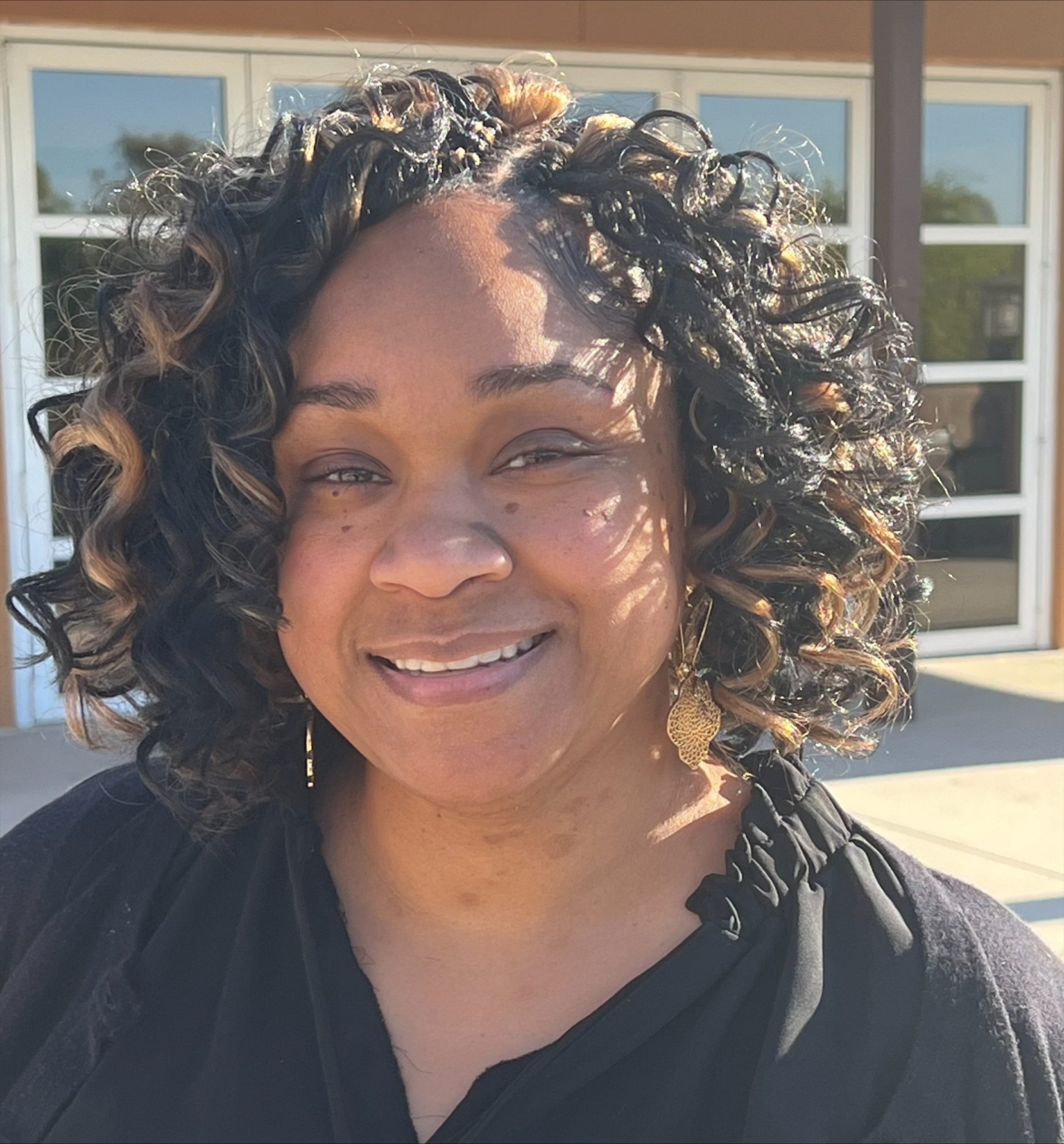 Picture of Karena Robertson a late 30s, lighter skinned African American woman with shoulder length blonde and black curly hair. Karena is smiling, wearing a black shirt with a building in the background.