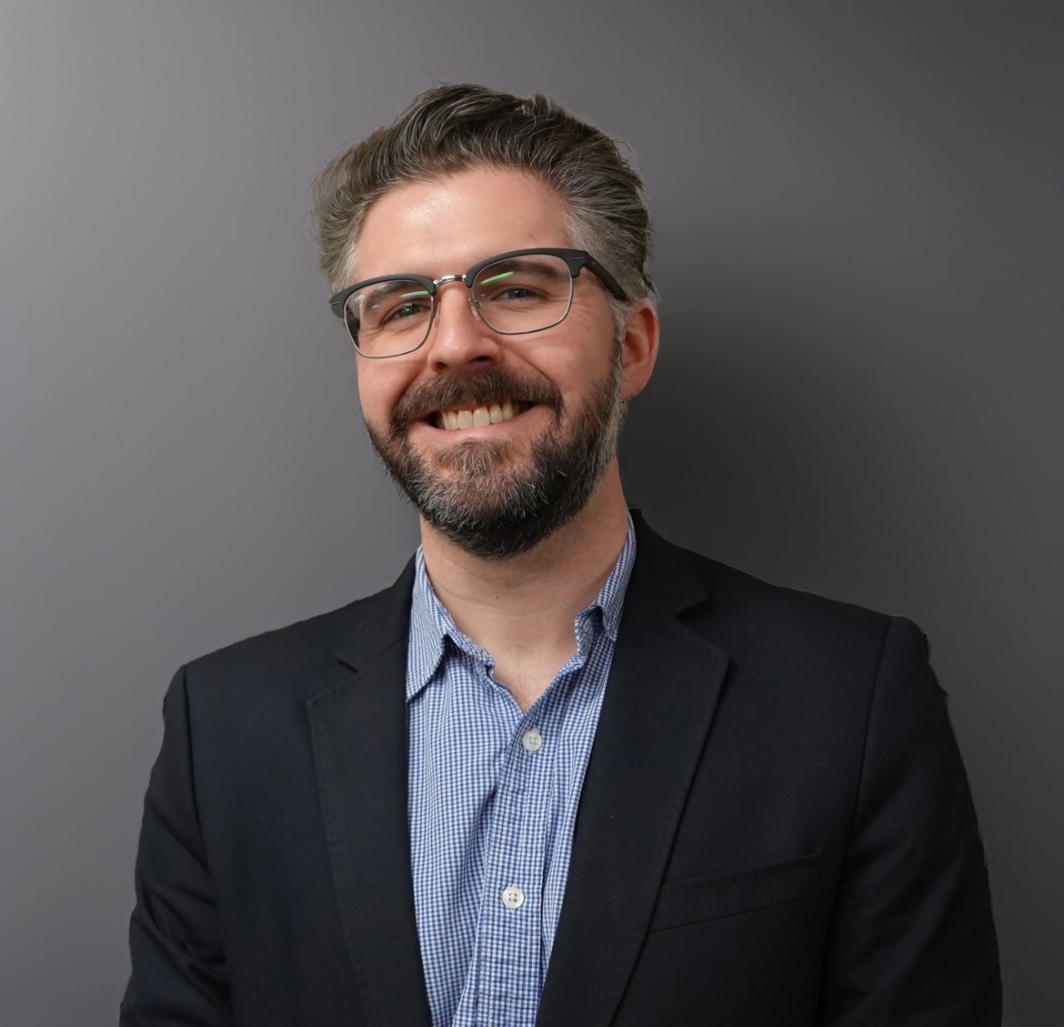 Andrew is a white male in his late 30s with dark and gray hair and short beard. He is wearing a blue blazer and blue shirt