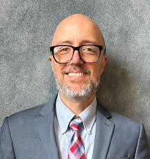 A man with a gray beard and black glasses smiles at the camera. He is standing in front of a solid gray background. He is wearing a light blue collared shirt with a red and blue tie and a gray suit jacket.