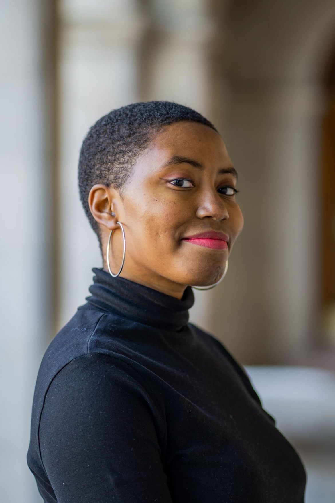 Olivia Williams, a young Black woman with low cut coily black hair wearing a black turtleneck, silver hoop earrings, and red lipstick. She is smiling softly at the camera.