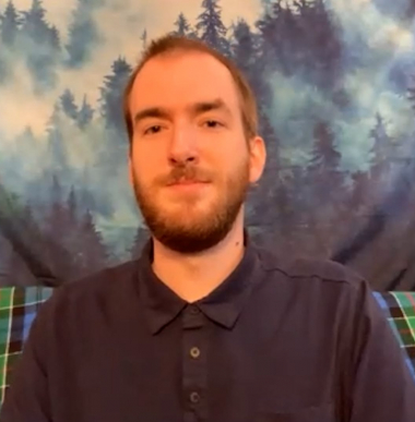 Christopher Hayes, a white male in his 20s with light brown heair and a beard, smiles directly at the camera. He is sitting infront of a tapestry hanging with forrest trees.