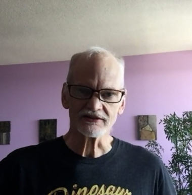 Crosby, an older white male wearing glasses, looks at the camera while in a room with lavendar walls