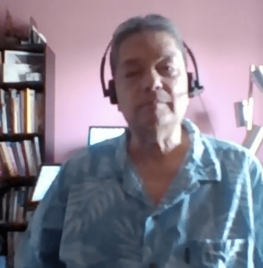 An asian american man sits in front of a pink wall wearing a floral shirt with a headset on