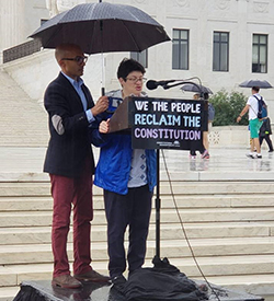Image of Liz Weintraub speaking at a podium in front of the Capitol Building in the rain. A man is standing next to her holding an umbrella over both of them..